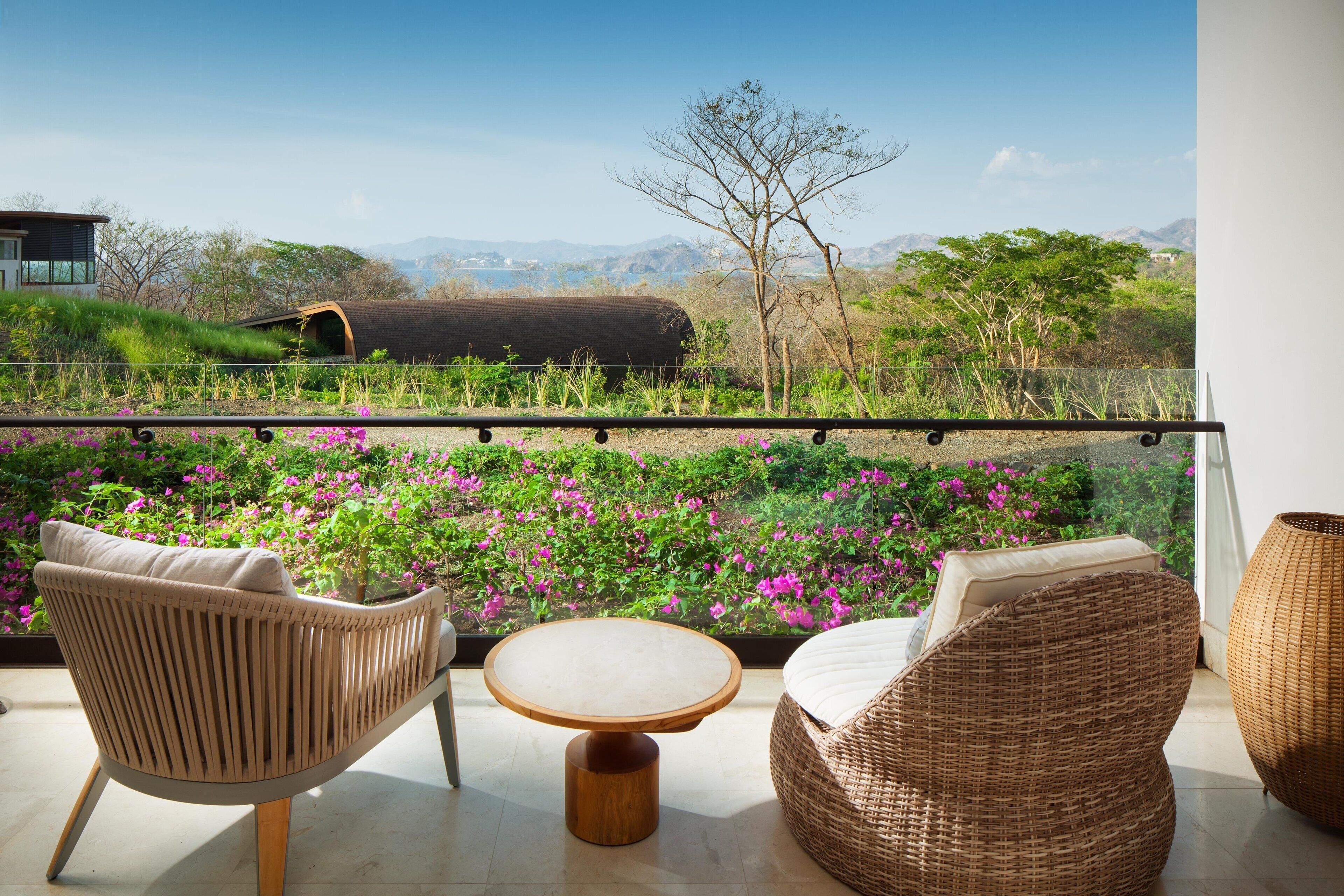 A balcony with wicker furniture, a round wooden table, and a view of a mountainous landscape with a train and greenery.