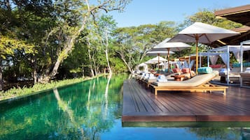 Zona junto a la piscina con sillas de descanso y sombrillas, rodeada de árboles.