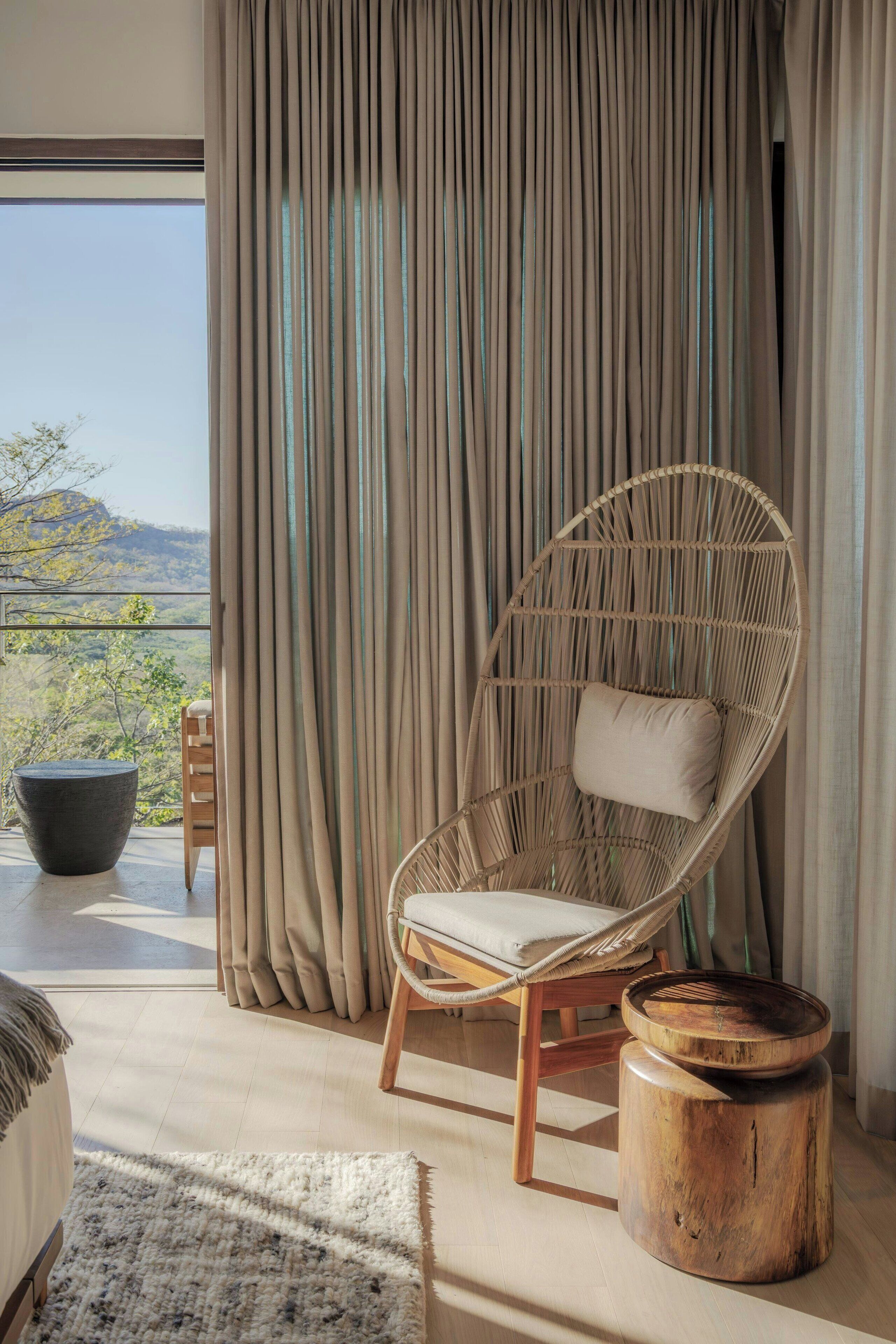 A modern room with a large window, a wicker chair, a wooden stool, and a view of the outdoors.