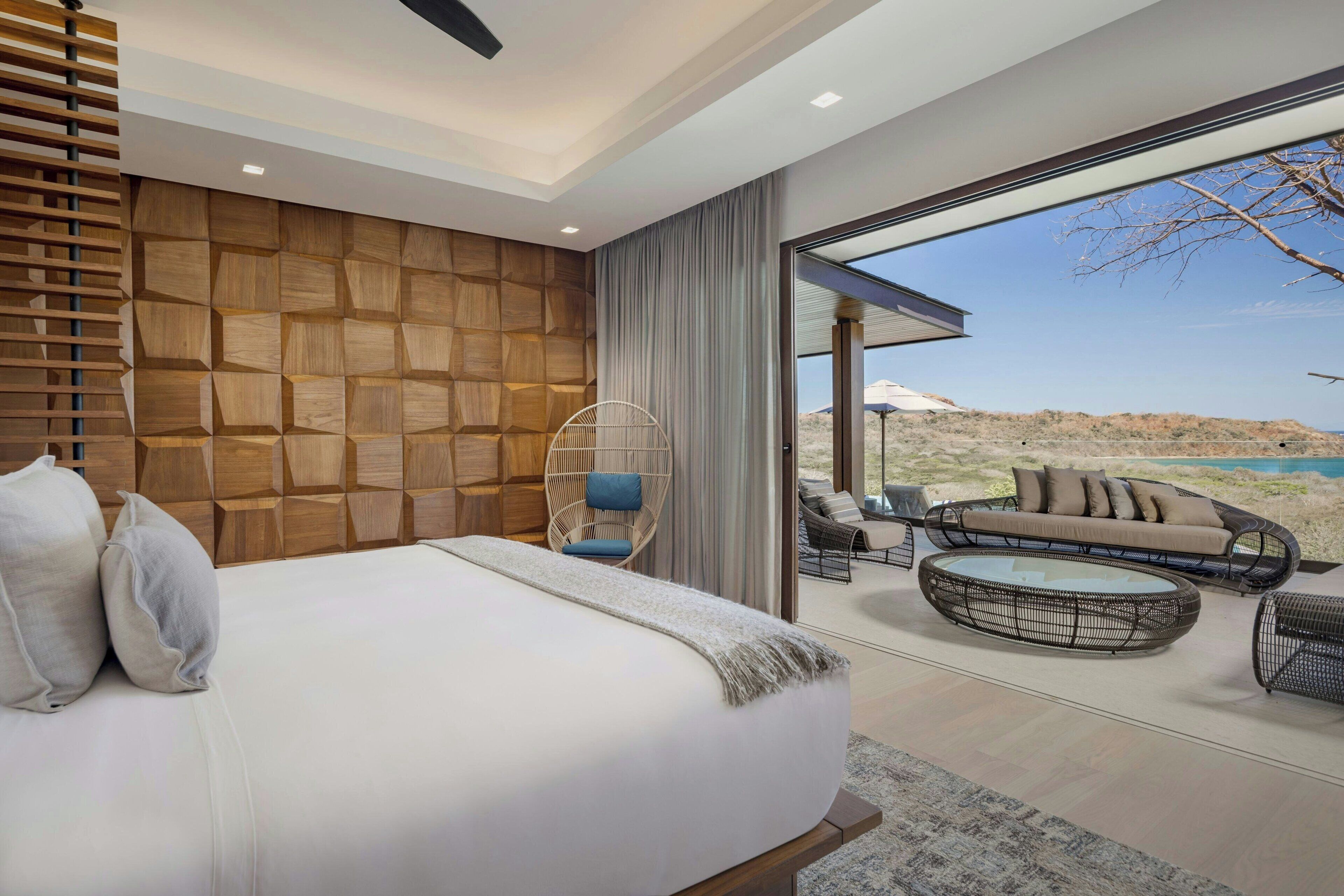 A modern bedroom with a large bed, a wooden wall panel, a wicker chair, a sofa, and a view of a desert landscape through a glass door.