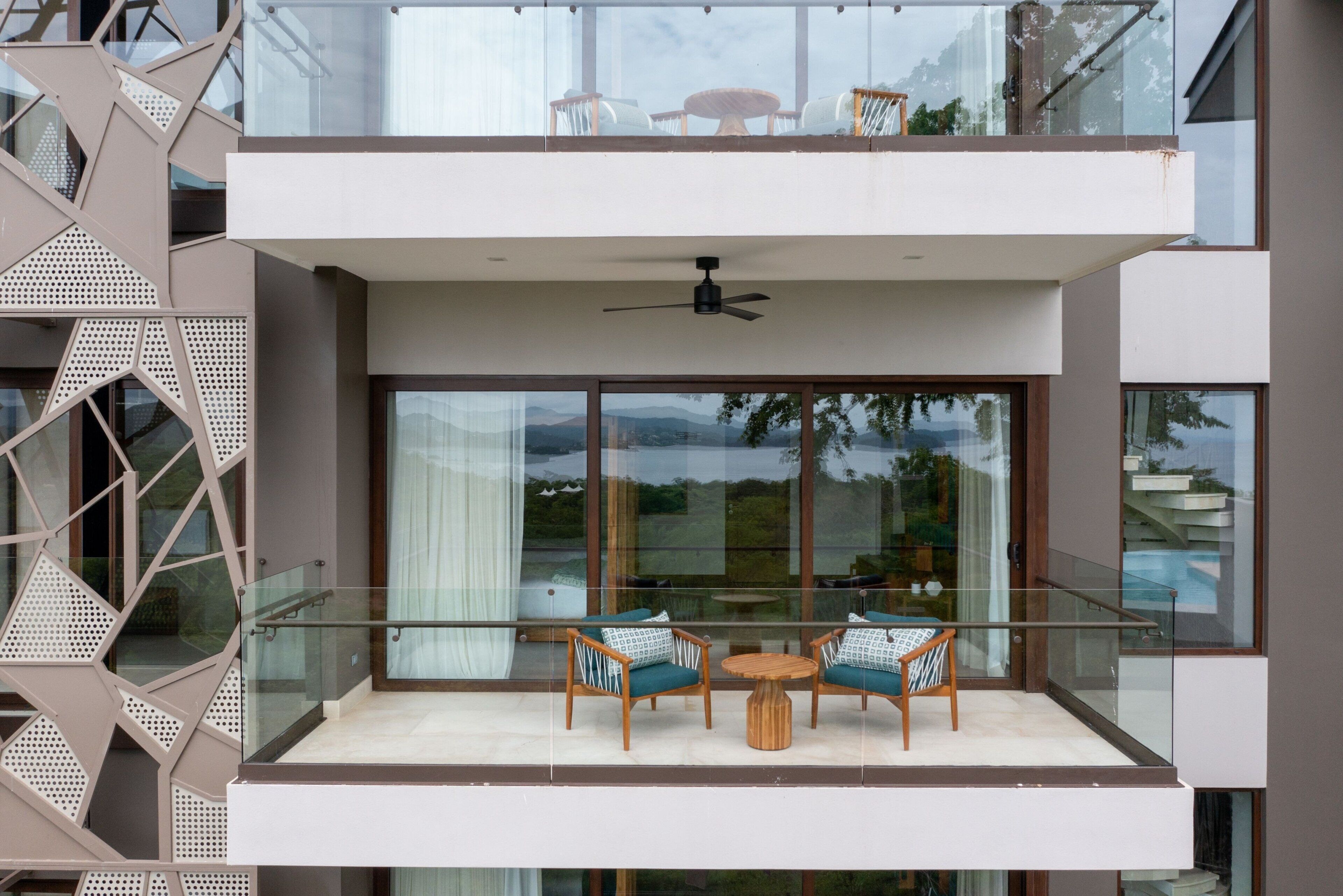 Een modern balkon met glazen hekwerk, twee houten stoelen, een klein rond tafeltje en een plafondventilator.