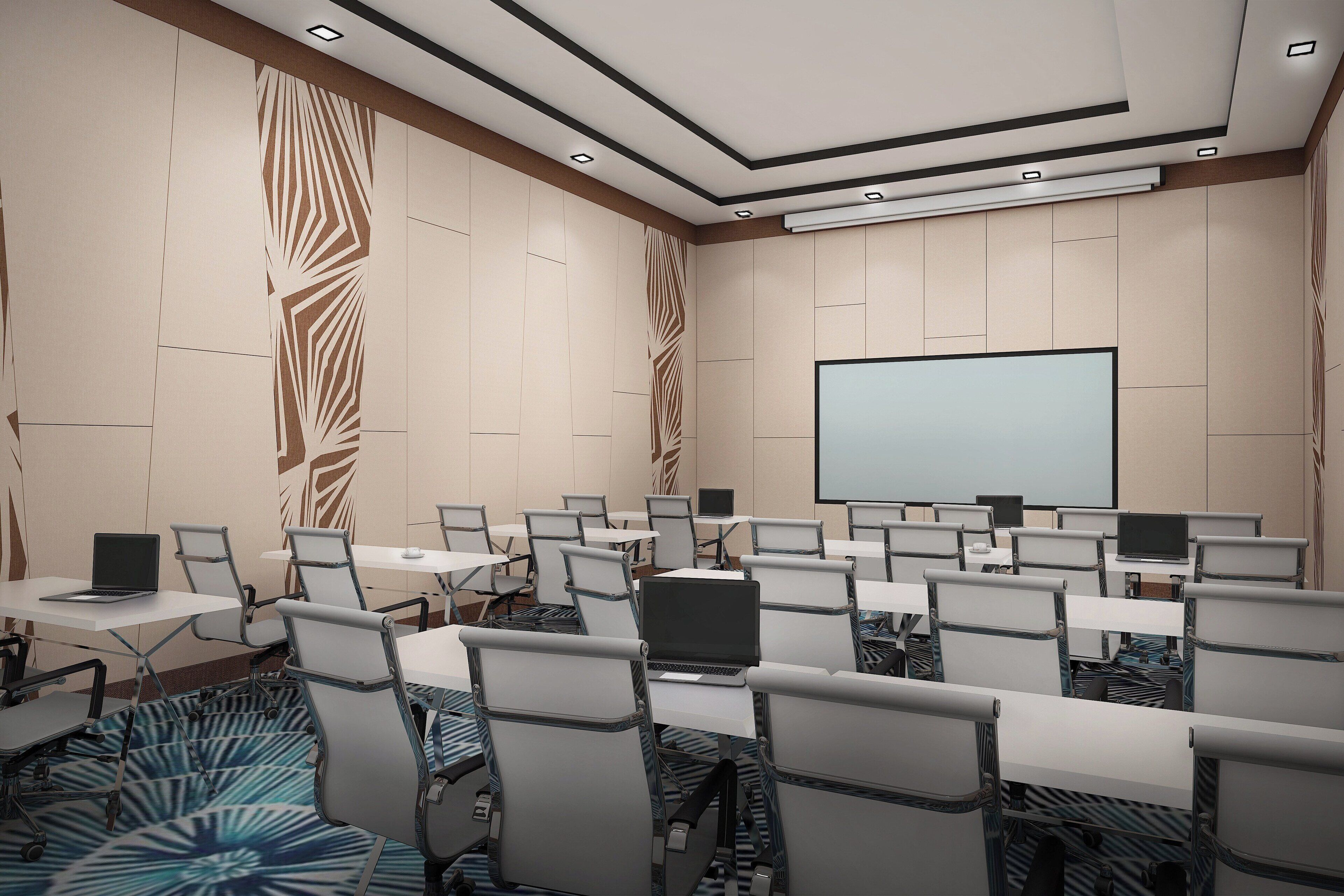 Un salón de conferencias con filas de mesas, sillas y portátiles, una pantalla de proyector y una alfombra estampada.