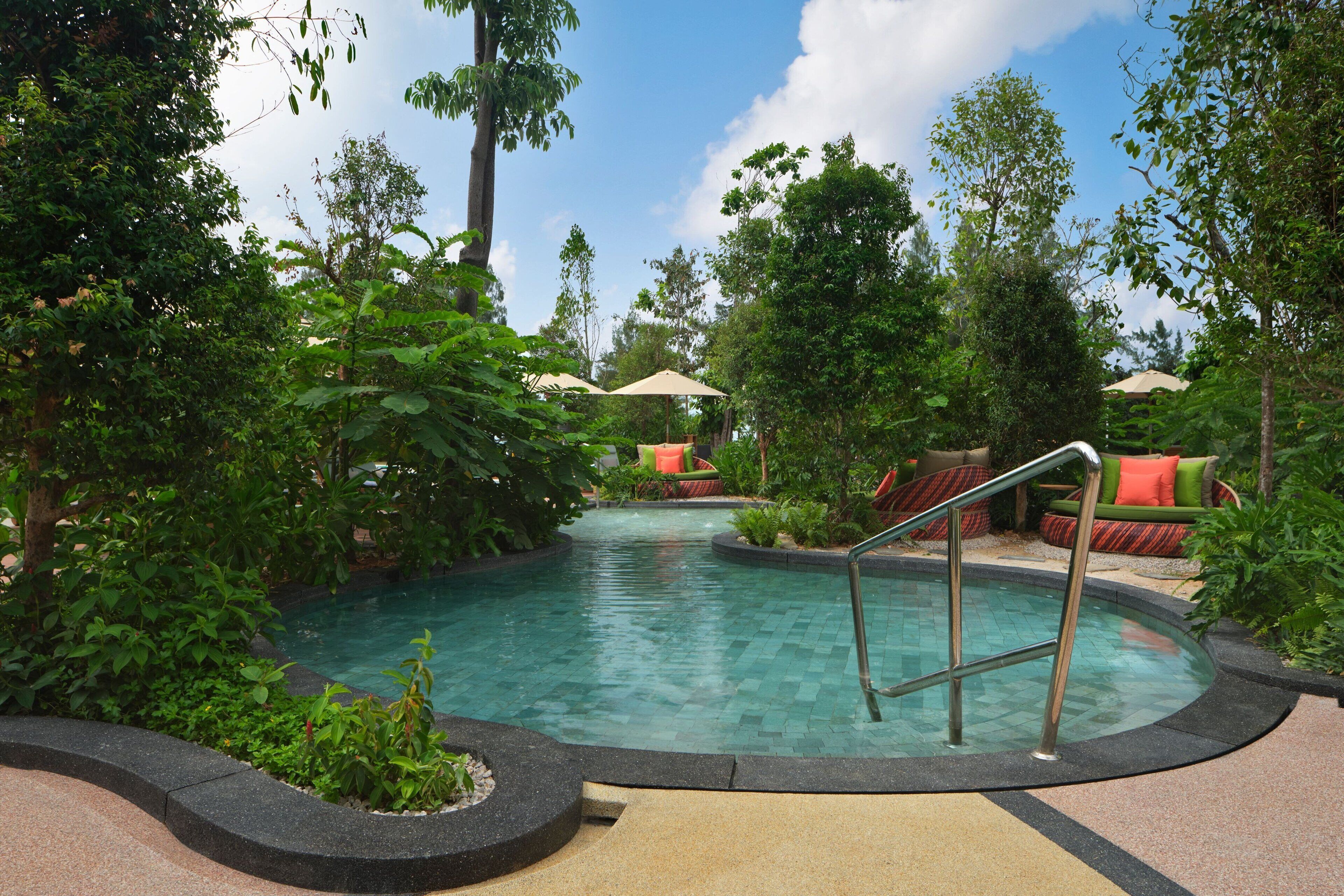 A pool area with a tiled deck, surrounded by lush greenery and outdoor seating with umbrellas.