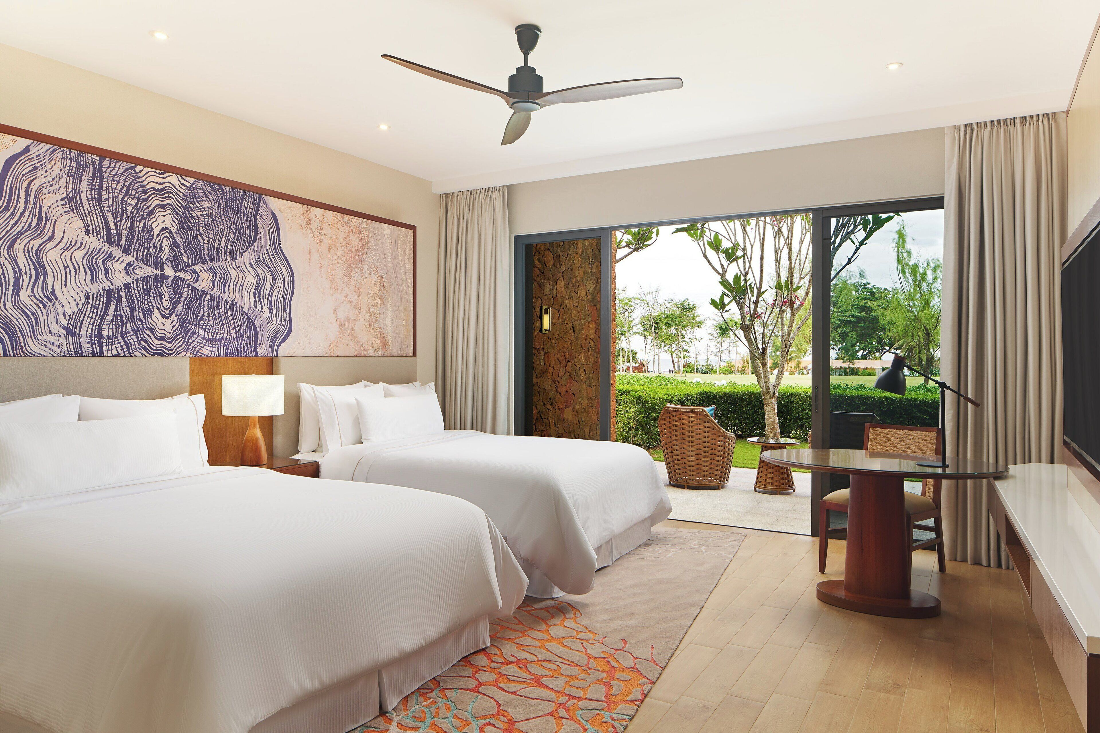 Habitación de hotel con dos camas, ventilador de techo, ventana grande y televisor.