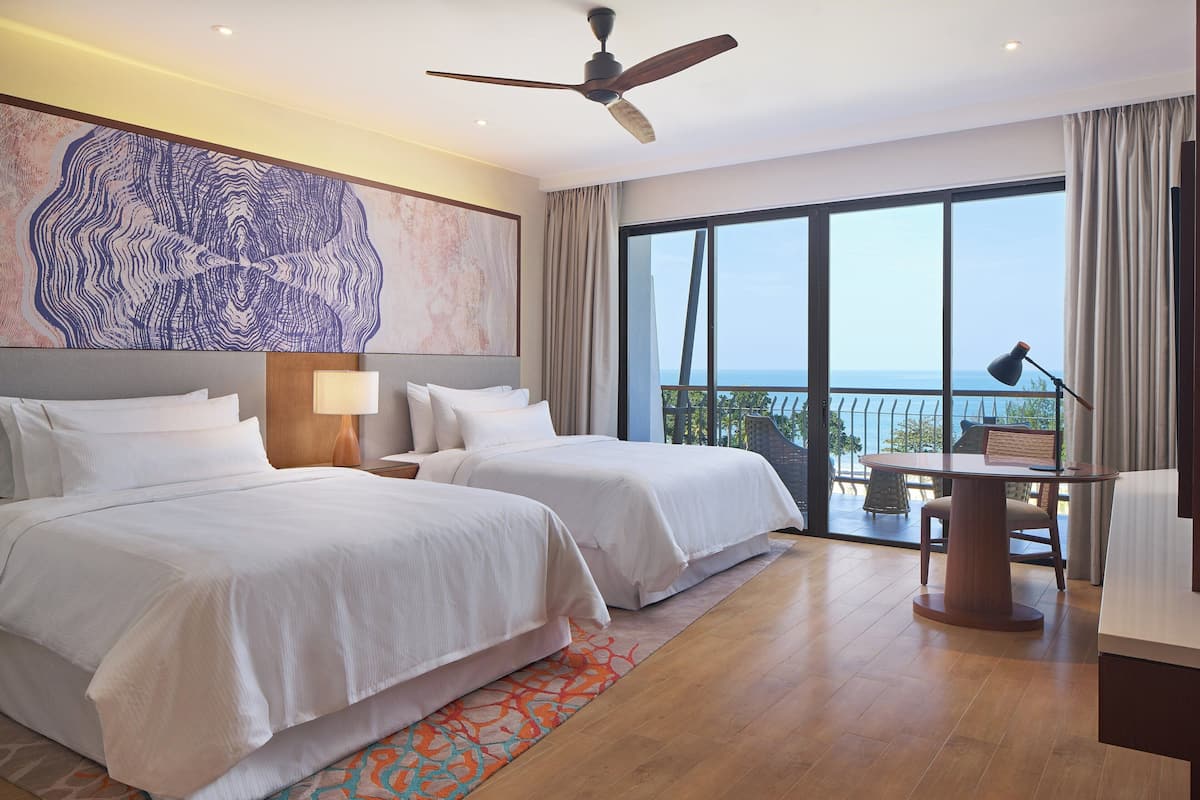 Ein Hotelzimmer mit zwei Betten, einem Deckenventilator, einem großen Fenster mit Blick und einem kleinen Tisch mit Lampe.