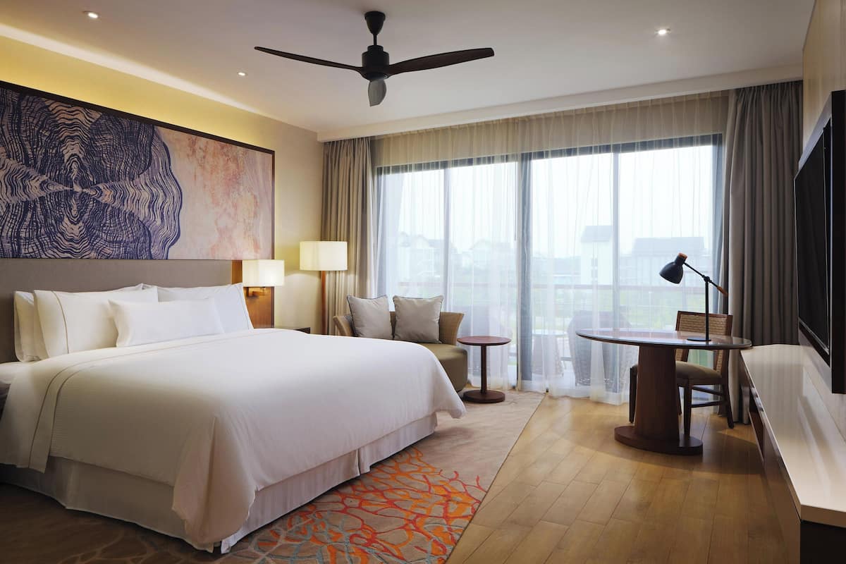 Ein Hotelzimmer mit einem großen Bett, einem Schreibtisch, einem Stuhl, einem Deckenventilator, einem Fernseher und einem großen Fenster mit Vorhängen.