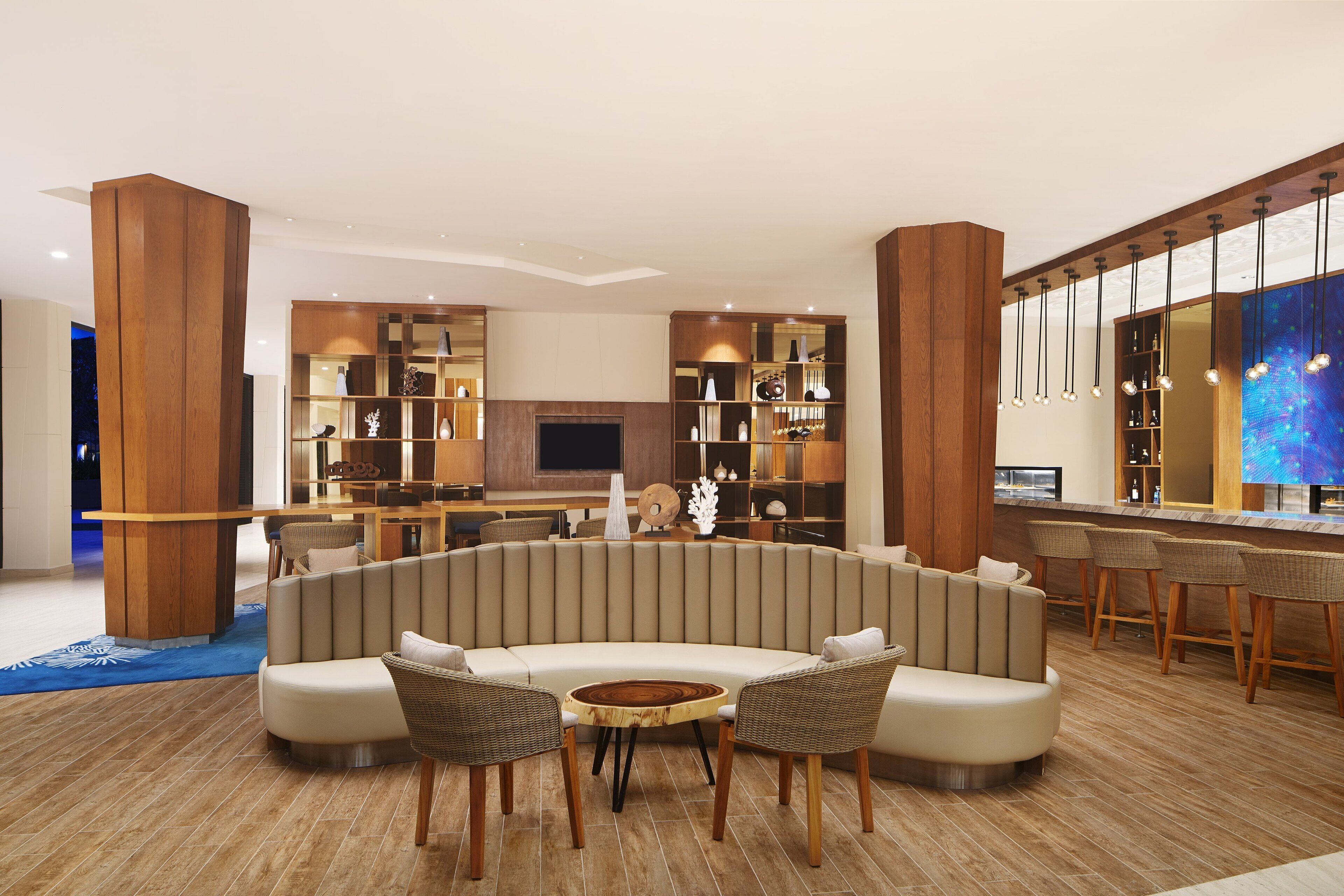 A modern lounge area with a curved sofa, a round wooden table, and wicker chairs. There is a TV mounted on the wall, a bar area with stools, and a large bookshelf filled with decorative items.