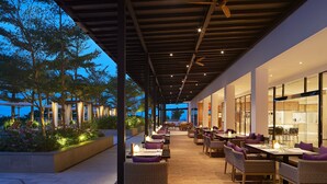 An outdoor dining area with tables and chairs, a covered walkway, and a view of trees and buildings. - The Westin Desaru Coast Resort (Bandar Penawar)