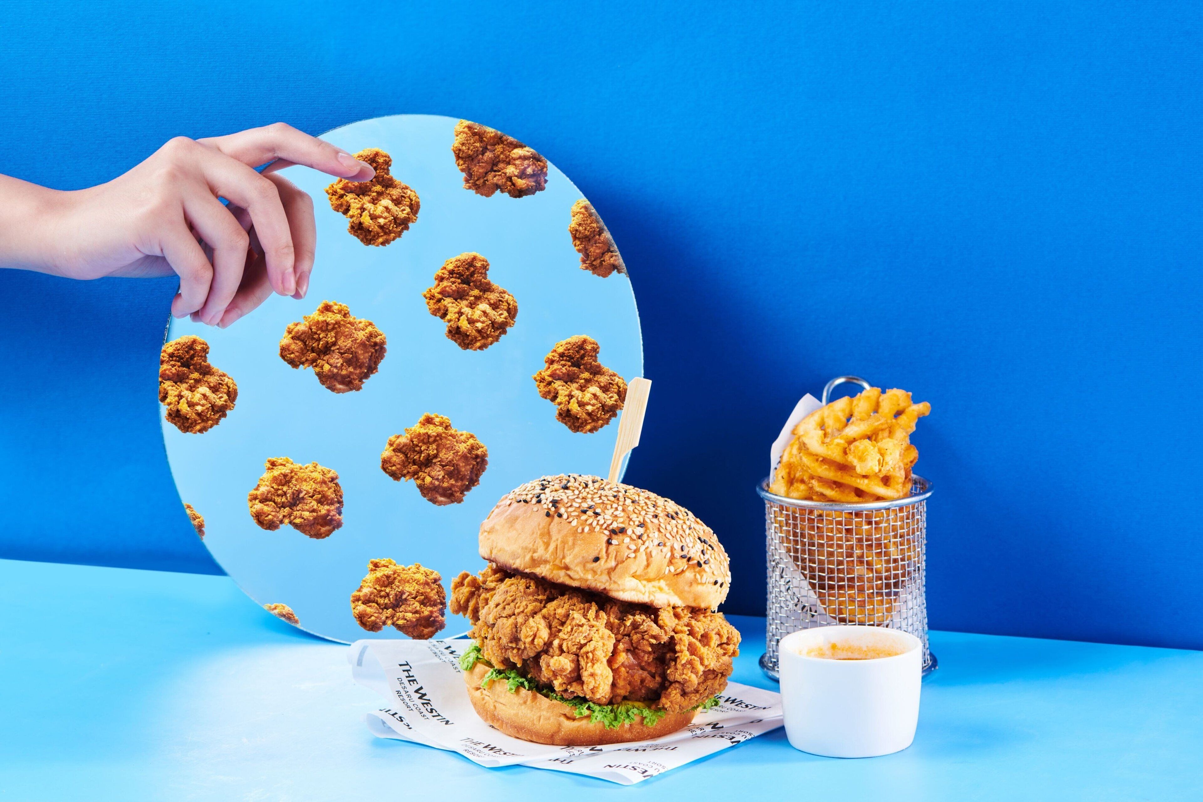 A hand holding a plate with fried chicken pieces, a burger, and a cup of fries on a blue surface.