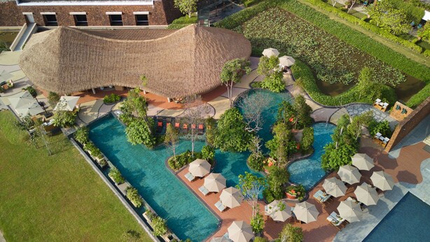 An aerial view of a resort with multiple swimming pools, a large thatched-roof structure, and numerous umbrellas and lounge chairs. - The Westin Desaru Coast Resort (Bandar Penawar)