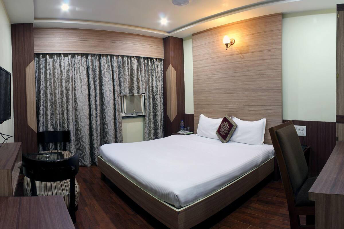 Quarto de hotel com uma cama grande, uma escrivaninha com cadeira, uma TV e uma janela com cortinas.