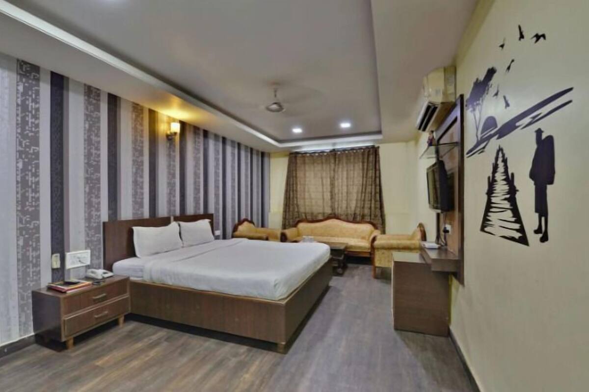 Quarto de hotel com uma cama grande, um sofá, uma TV fixada na parede e uma cortina na janela.