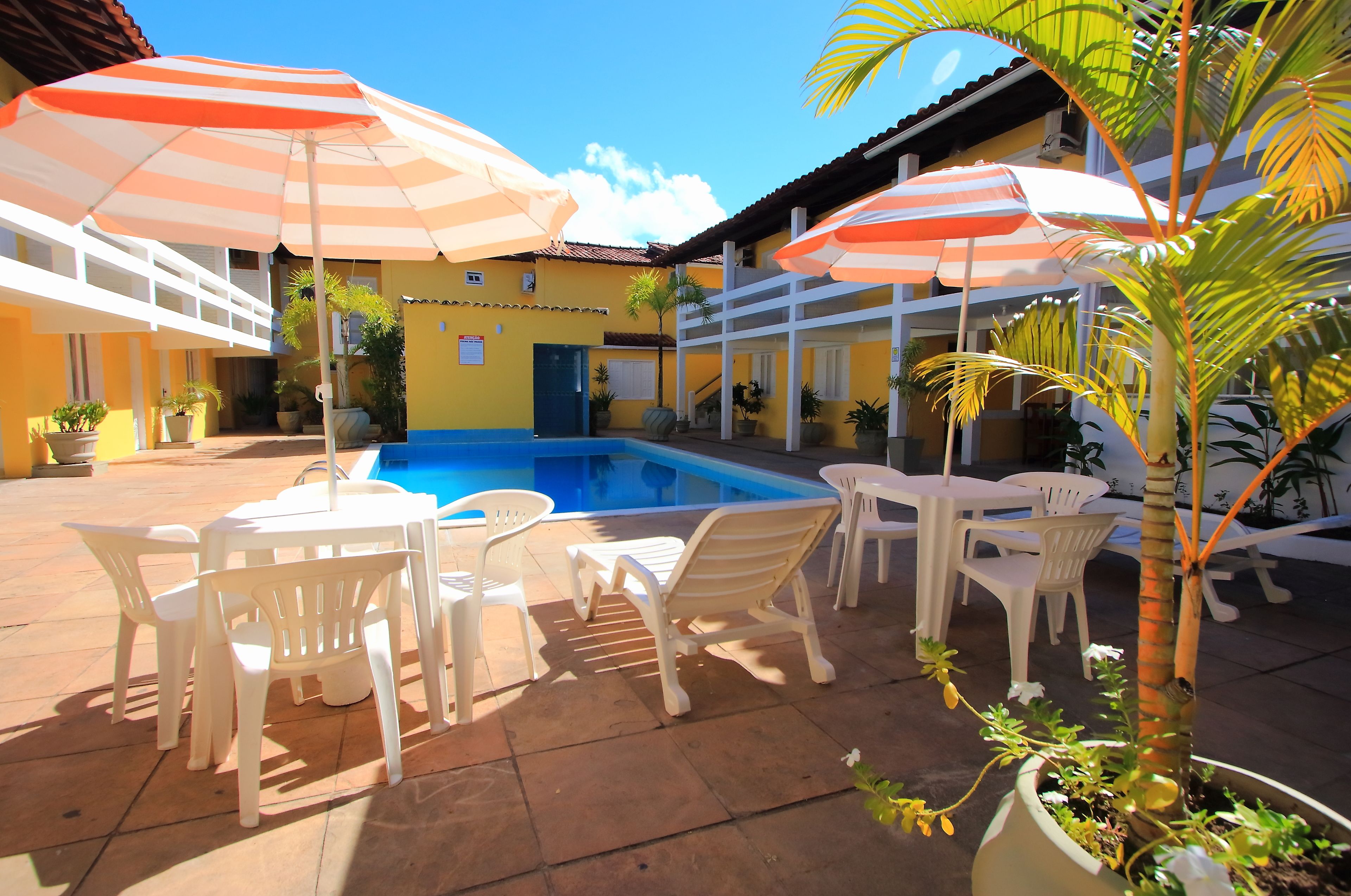 Zona junto a la piscina con sillas y mesas de plástico blanco, sombrillas a rayas y plantas en macetas.