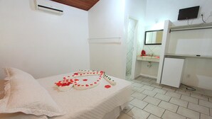 A bedroom with a bed, a small table with a decorative item, a white wall, a door, a mirror, and a television on the wall. - Rede Andrade Porto Seguro Navegantes (Porto Seguro)