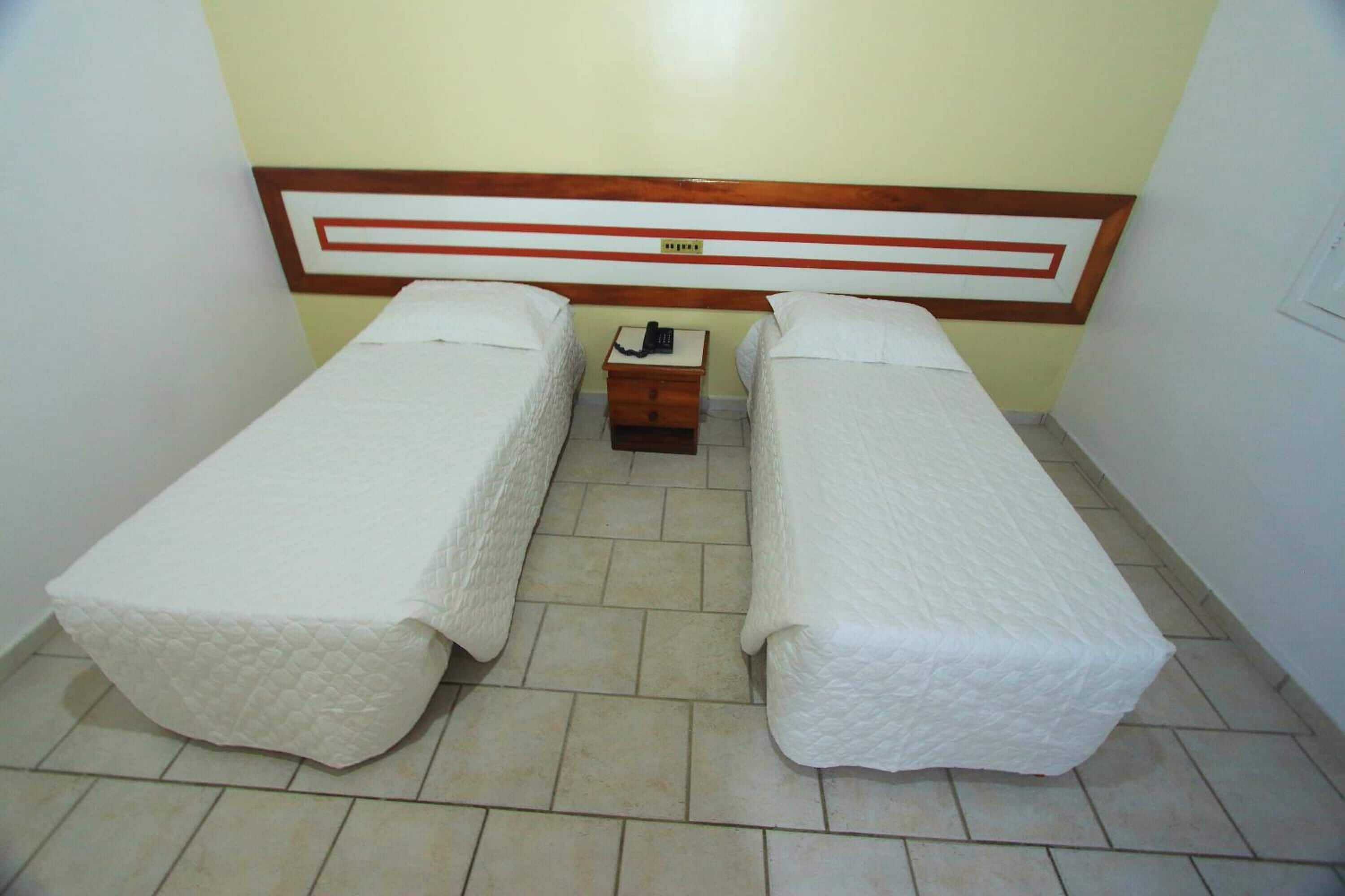 A small, clean room with two single beds, a wooden headboard, a bedside table with a phone, and a tiled floor.