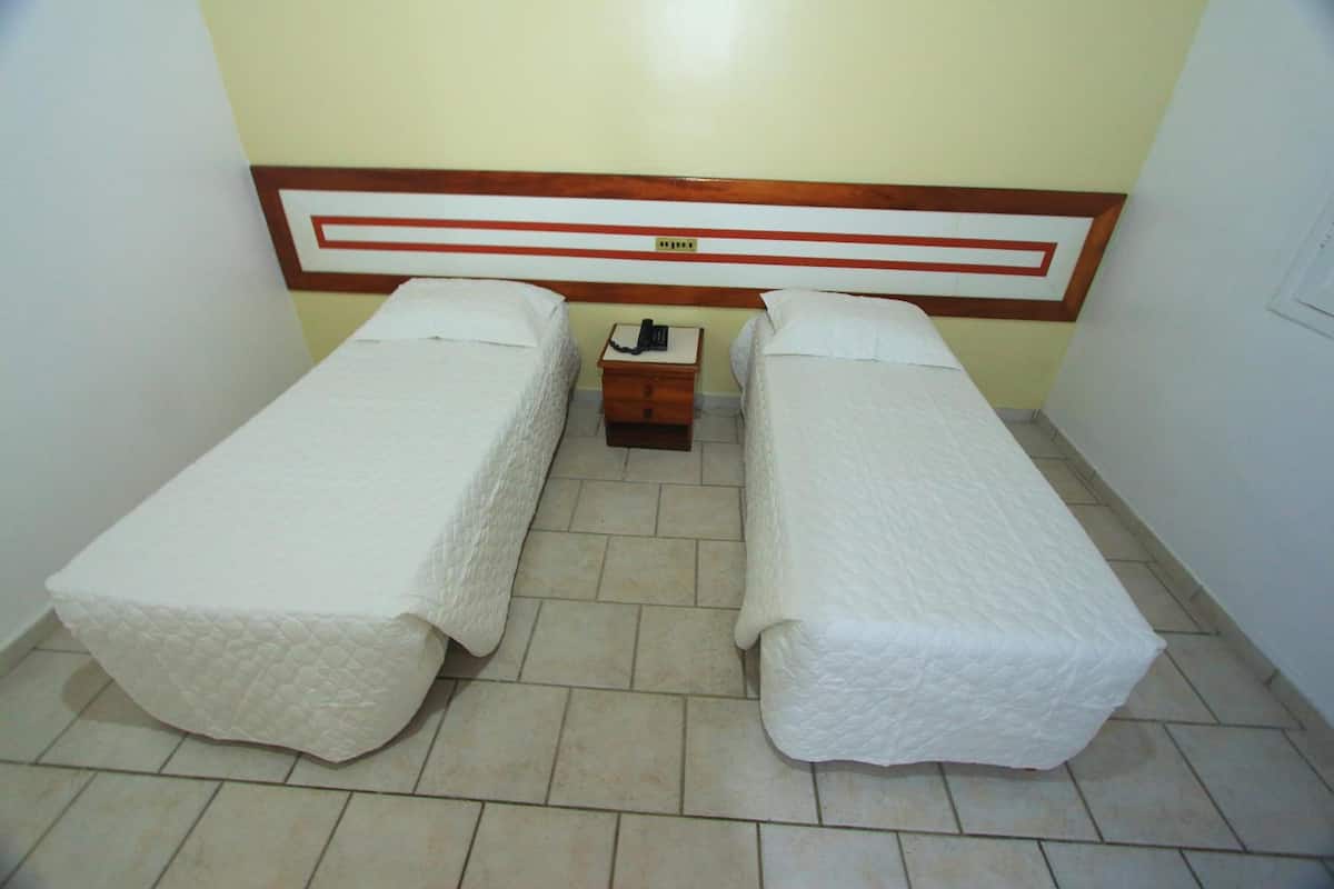 Una habitación pequeña y limpia con dos camas individuales, un cabecero de madera, una mesita de noche con teléfono y suelo de baldosas.