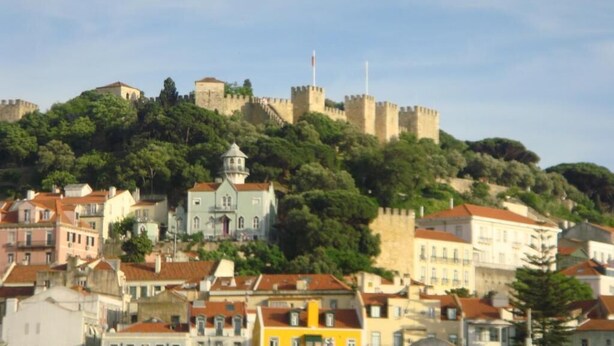 A historic cityscape with a prominent castle on a hill, surrounded by colorful buildings and lush greenery. - MIDIS - Local Accommodation (Lisbon)