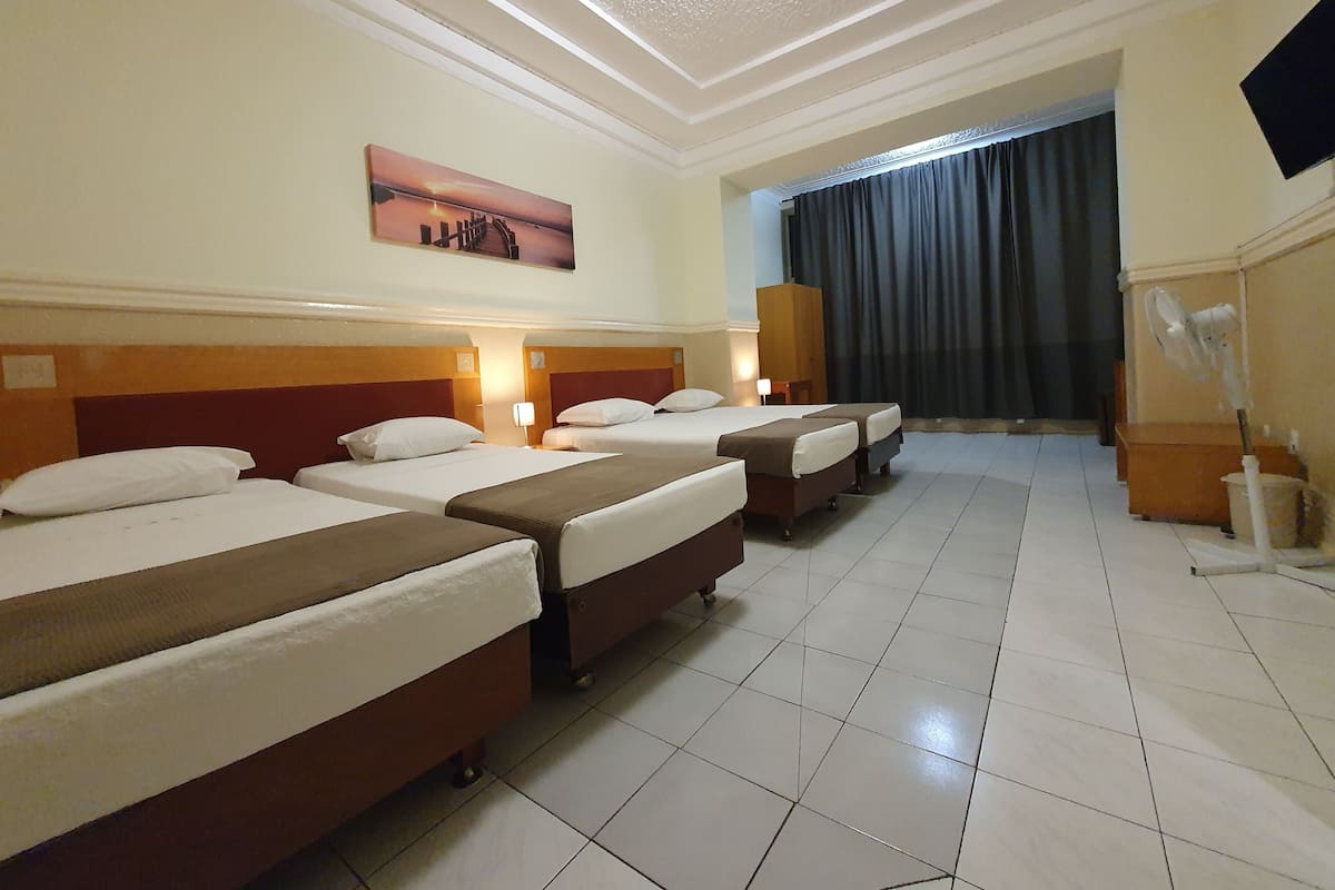 A hotel room with four beds arranged in a row, a painting on the wall, a large window with curtains, and a television mounted on the wall.
