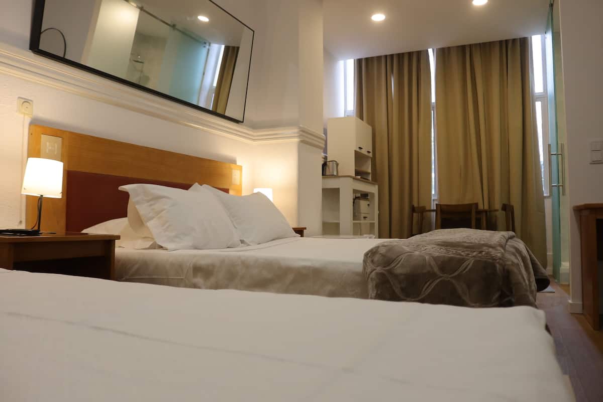 A hotel room with two beds, a wooden headboard, a bedside table with a lamp, a mirror, a wardrobe, and a window with curtains.