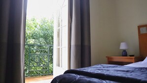 A bedroom with a bed, a nightstand with a lamp, and a view of greenery through a window. - MIDIS - Local Accommodation (Lisbon)