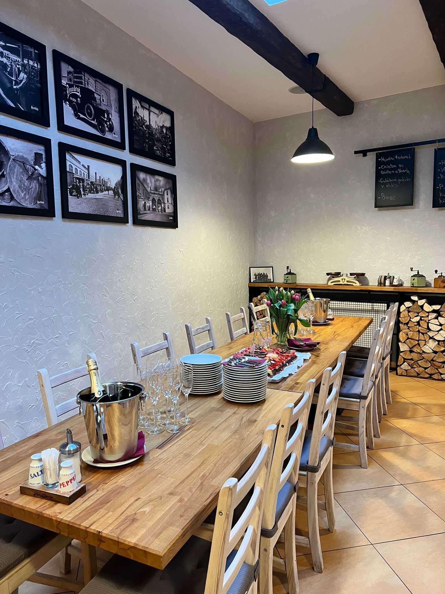 A dining area with a wooden table set for a meal, surrounded by chairs. There are framed black and white photographs on the wall, a chalkboard menu, and a wooden shelf with kitchen items.