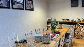 A dining area with a wooden table set for a meal, surrounded by chairs. There are framed black and white photographs on the wall, a chalkboard menu, and a wooden shelf with kitchen items.
