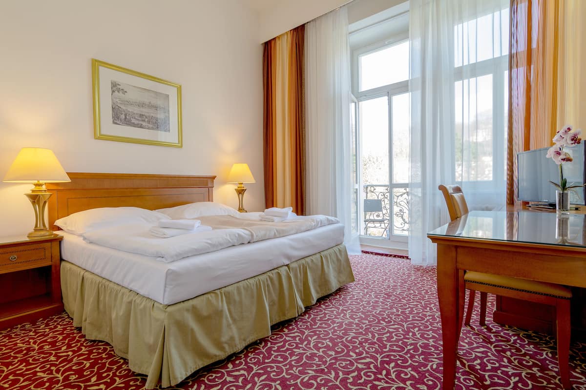 A hotel room with a neatly made bed, two bedside lamps, a large window with curtains, a wooden desk, and a framed picture on the wall.
