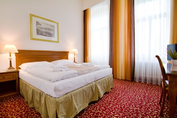 A neatly made bed with white linens, a wooden headboard, and a red patterned carpet. There are two bedside lamps and a framed picture on the wall. - Hotel Romania (Karlovy Vary)