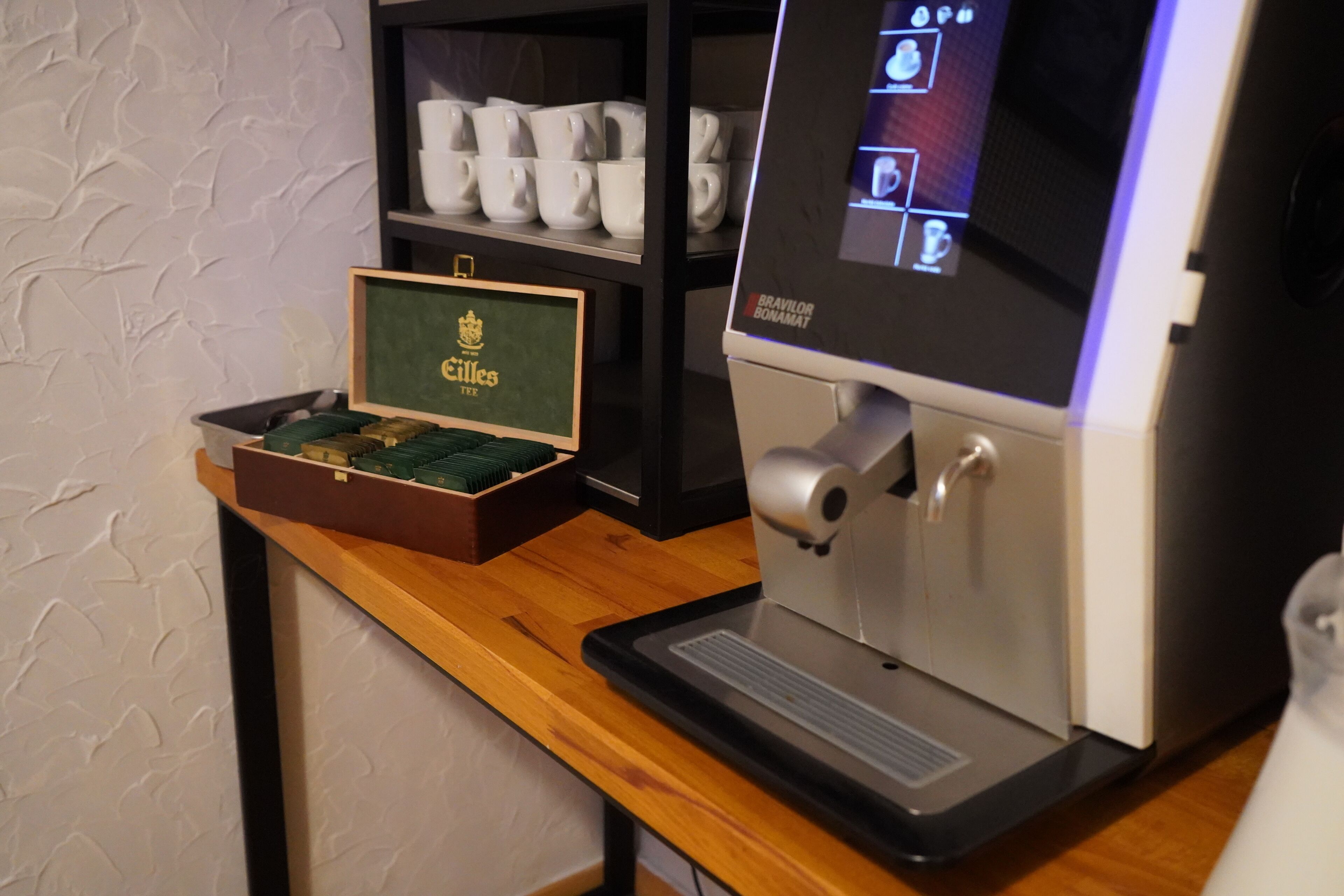 A coffee machine with a digital display, a coffee cup holder, and a box of coffee beans on a wooden counter.