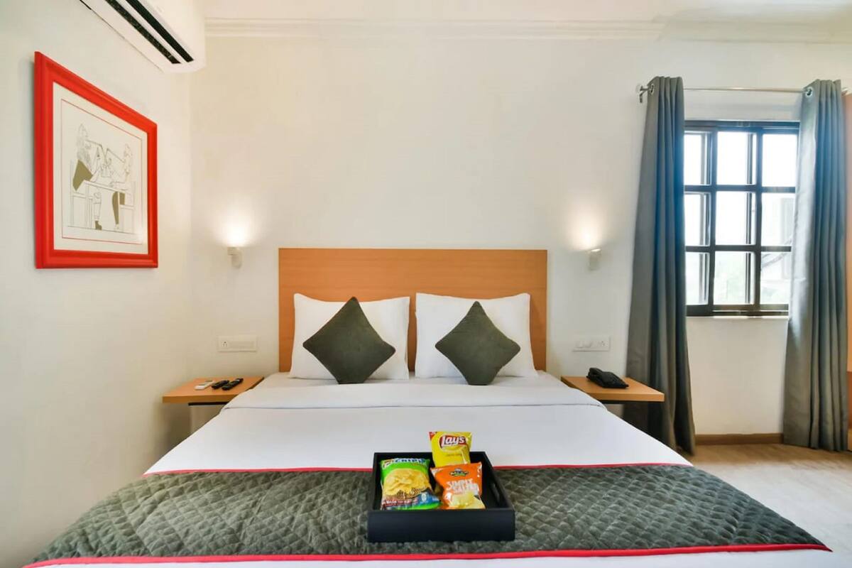 A hotel room with a bed, two pillows, a bedside table with a phone, a window with curtains, and a wall with a framed picture.