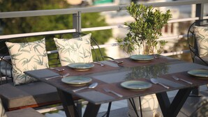 Mesa de comedor preparada con platos, cubiertos y una planta en maceta en un balcón con mobiliario exterior.