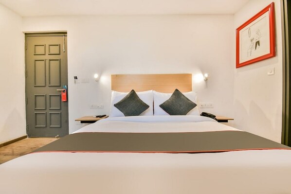 A hotel room with a bed, a nightstand, a bedside table, a door, and a framed picture on the wall. - Hotel Broadway Udaipur (Udaipur)