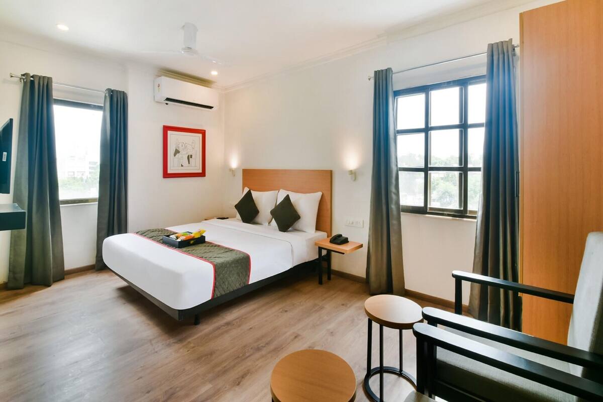 A hotel room with a bed, bedside table, nightstand, and a small seating area with a chair and stool.