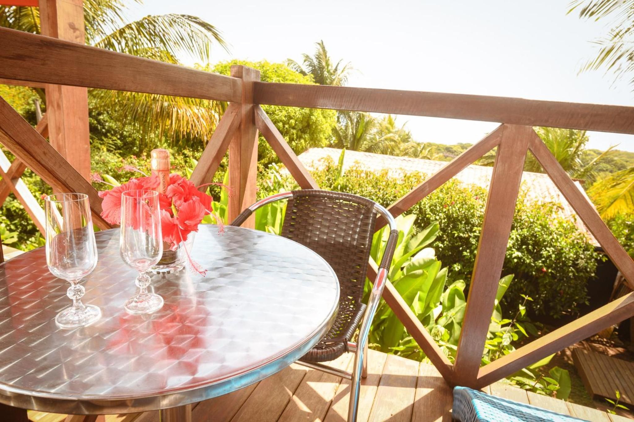 Uma mesa de vidro redonda com duas taças e uma garrafa, uma cadeira de vime, e um deck de madeira com vista para vegetação.