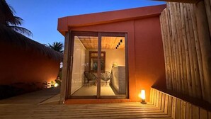 Un bâtiment moderne doté d’une porte en verre, d’une terrasse en bois et, au fond, d’une structure à toit de chaume.