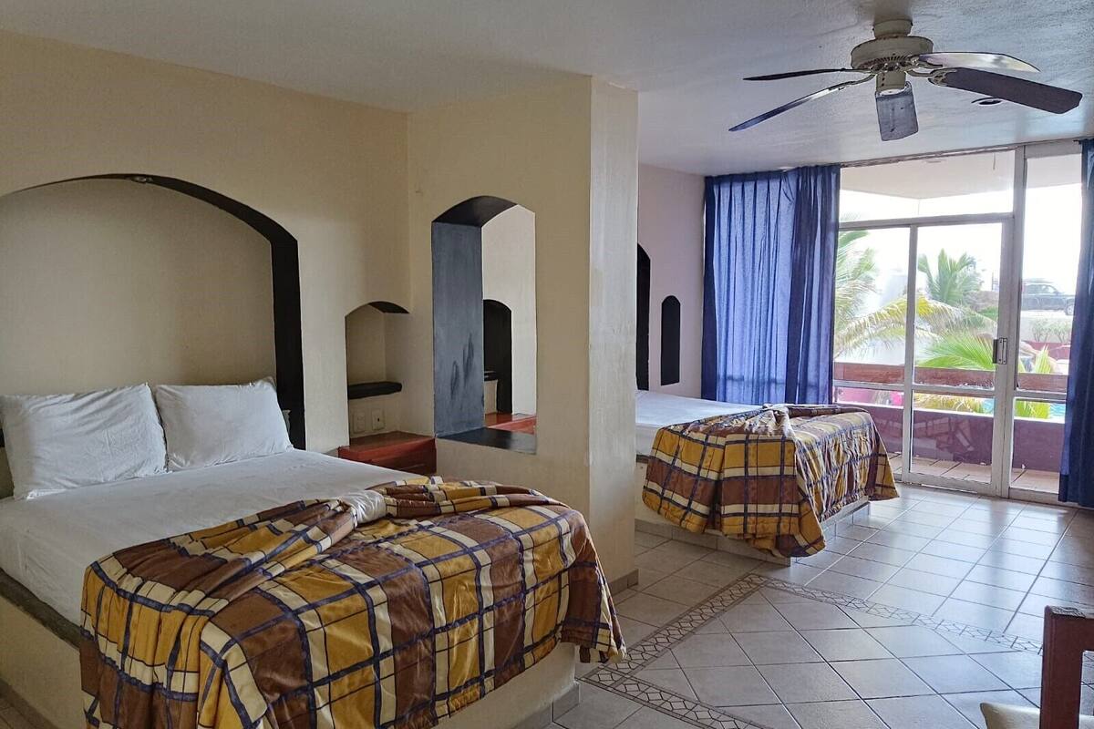 A hotel room with two beds, a ceiling fan, a balcony with curtains, and a view of the outdoors.