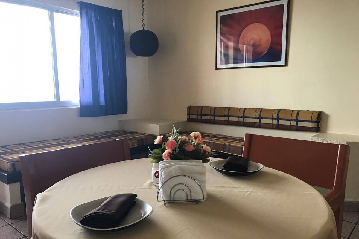 A dining area with a round table, chairs, a vase with flowers, a hanging light, and a framed picture on the wall.