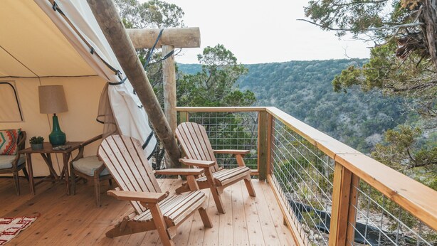 A wooden deck with Adirondack chairs, a table with a lamp, and a view of a forested landscape. - Collective Hill Country Retreat (Wimberley)