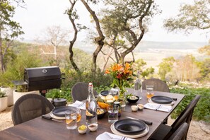 A dining table set outdoors with a grill, chairs, and a view of trees and hills. - Collective Hill Country Retreat (Wimberley)