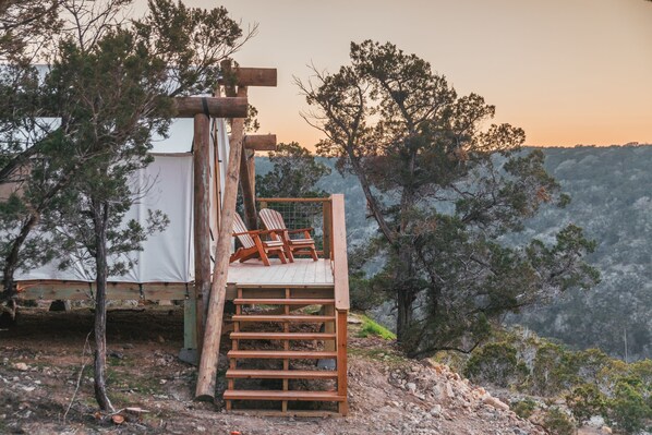 A wooden deck with a staircase leading to a bed area, surrounded by trees and a tent in a natural setting. - Collective Hill Country Retreat (Wimberley)