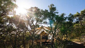 A wooden cabin surrounded by trees with a clear sky and sunlight filtering through. - Collective Hill Country Retreat (Wimberley)