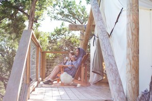 A wooden deck with two people relaxing in chairs, a white tent, and surrounding trees. - Collective Hill Country Retreat (Wimberley)
