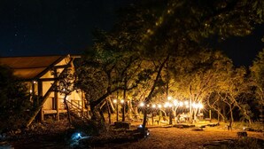 A rustic outdoor setting with a wooden structure, possibly a cabin, surrounded by trees and illuminated by string lights. - Collective Hill Country Retreat (Wimberley)