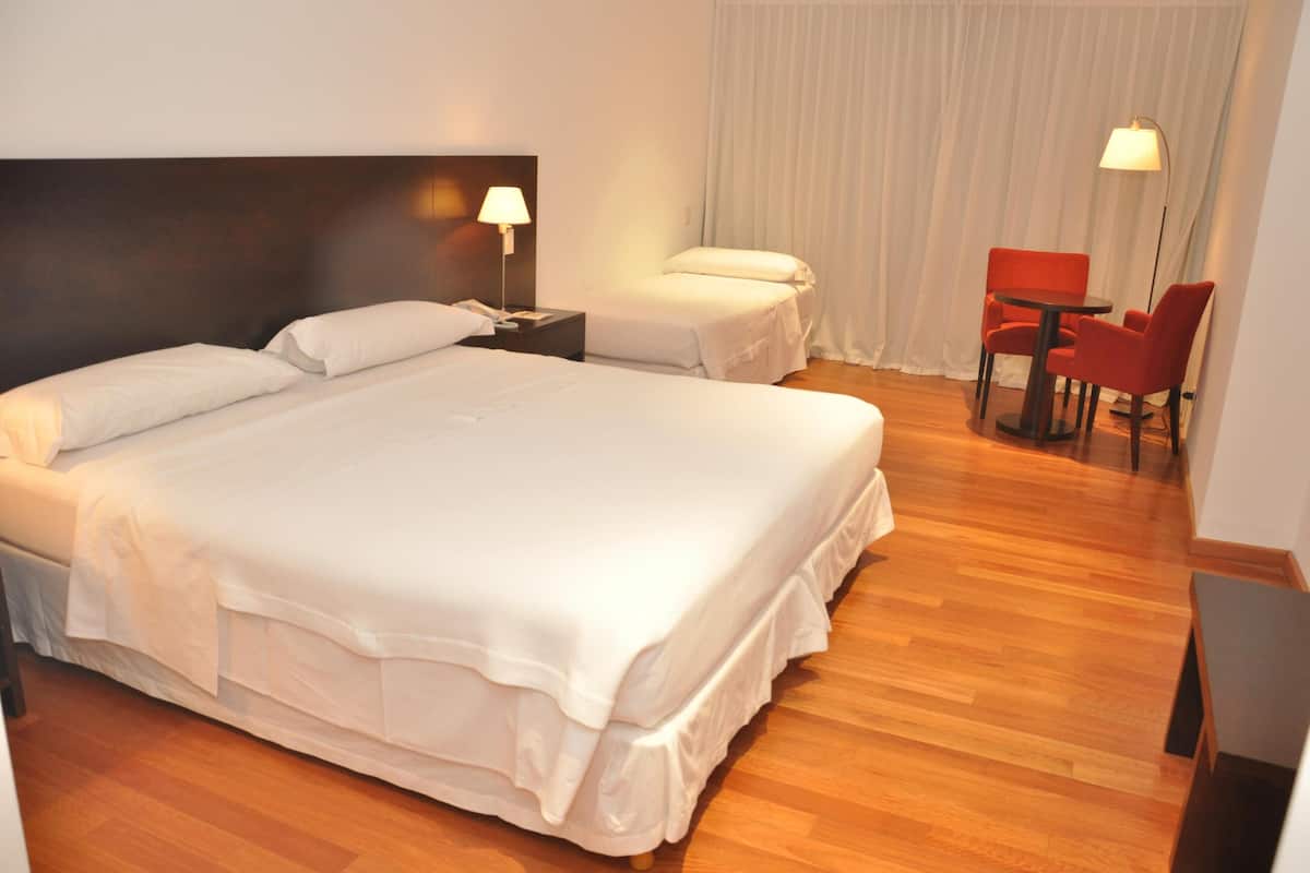 A hotel room with a large bed, a bedside table with a lamp, a separate bed, a chair, and a table.