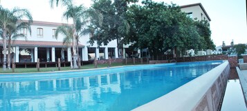 Una piscina con borde de baldosas, rodeada de palmeras y un edificio con techo de tejas.