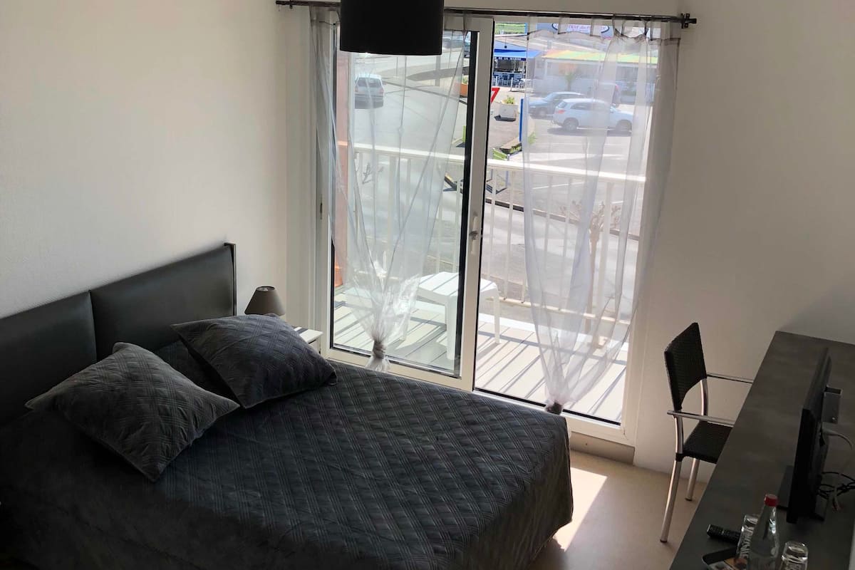 A bedroom with a bed, a desk, a chair, and a large window with a view of the street.