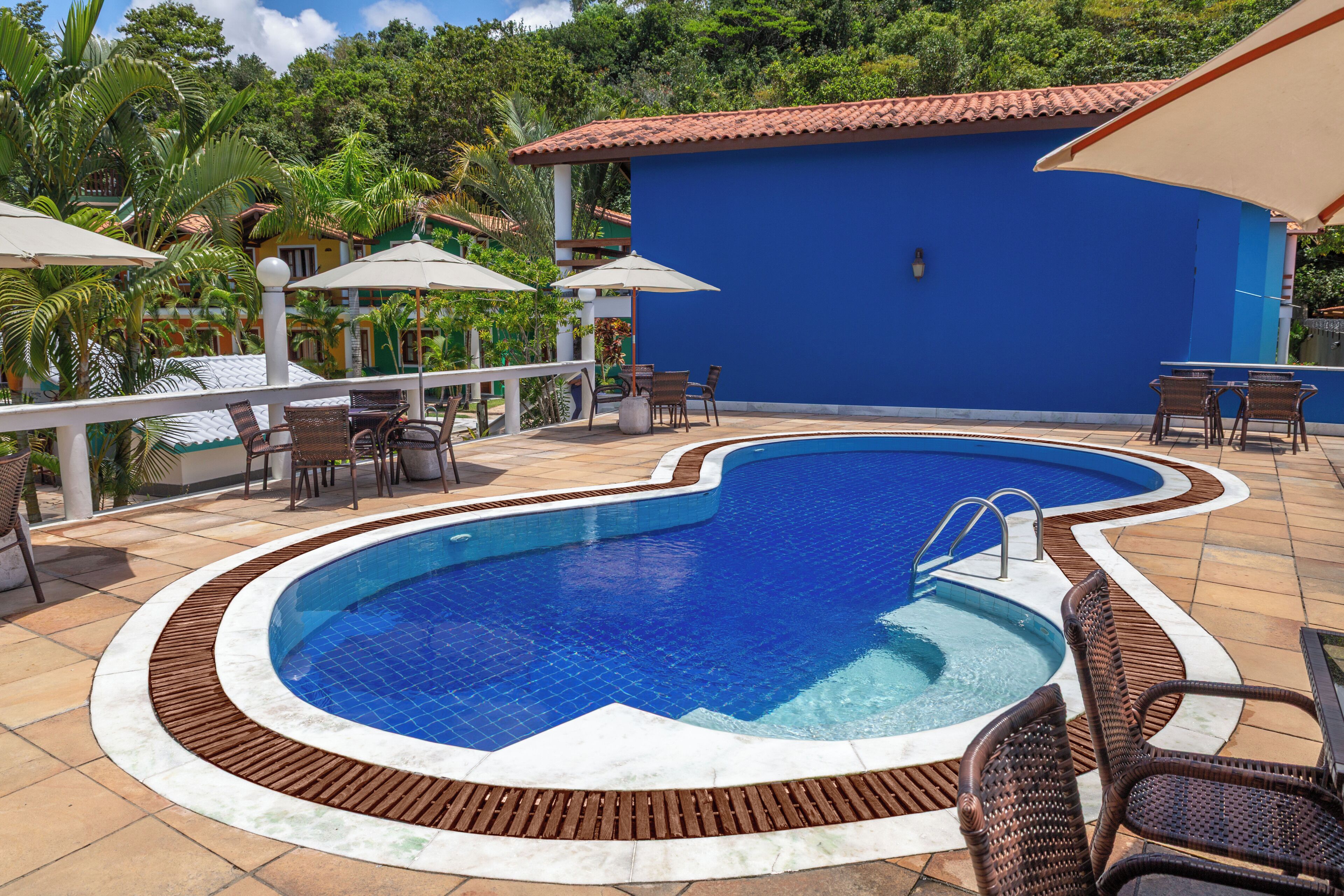 A swimming pool with a tiled edge, surrounded by lounge chairs and umbrellas, set against a blue building with a tiled roof.