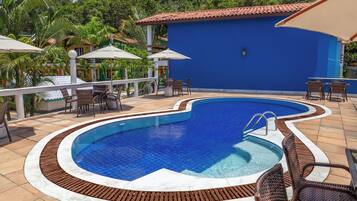 A swimming pool with a tiled edge, surrounded by lounge chairs and umbrellas, set against a blue building with a tiled roof.