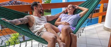 Een hangmat tussen twee personen op een balkon met een betegelde vloer en uitzicht op een blauw gebouw.