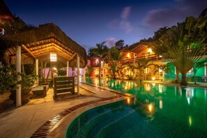 A swimming pool with a thatched-roof structure and a building with balconies. - Capoeira Village (Porto Seguro)
